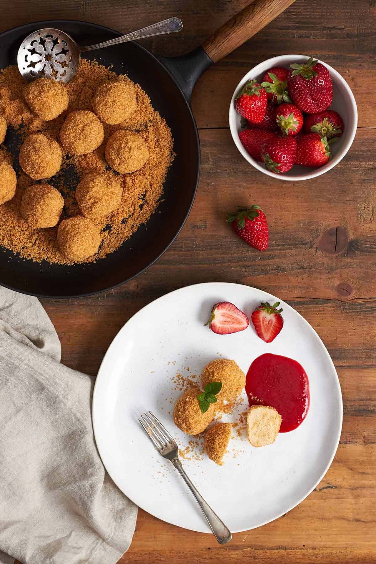 von oben: drei topfenknödel mit butterbröseln auf weißem teller, einer davon durchgeschnitten und mit gabel aufgespiest. stellfläche aus holz. oberhalb topfenknödel und butterbrösel in einer gußeisenen pfanne und und eine weiße schüssel mit frischen erdbeeren.