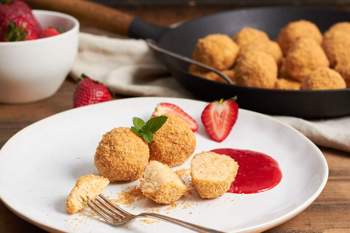 drei topfenknödel mit butterbröseln auf weißem teller, einer davon durchgeschnitten und mit gabel aufgespiest. stellfläche aus holz. im hintergrund topfenknödel und butterbrösel in einer gußeisenen pfanne und eine durchsichtige schüssel mit erdbeersauce.