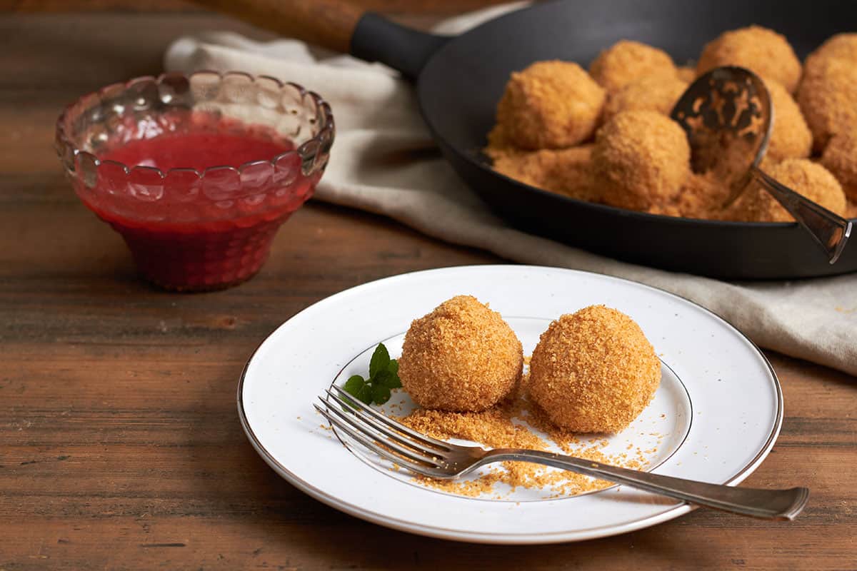 zwei topfenknödel mit butterbröseln und eine gable auf einem weißen teller und stellfläche aus holz. im hintergrund topfenknödel und butterbrösel in einer gußeisene pfanne und eine durchsichtige schüssel mit erdbeersauce.