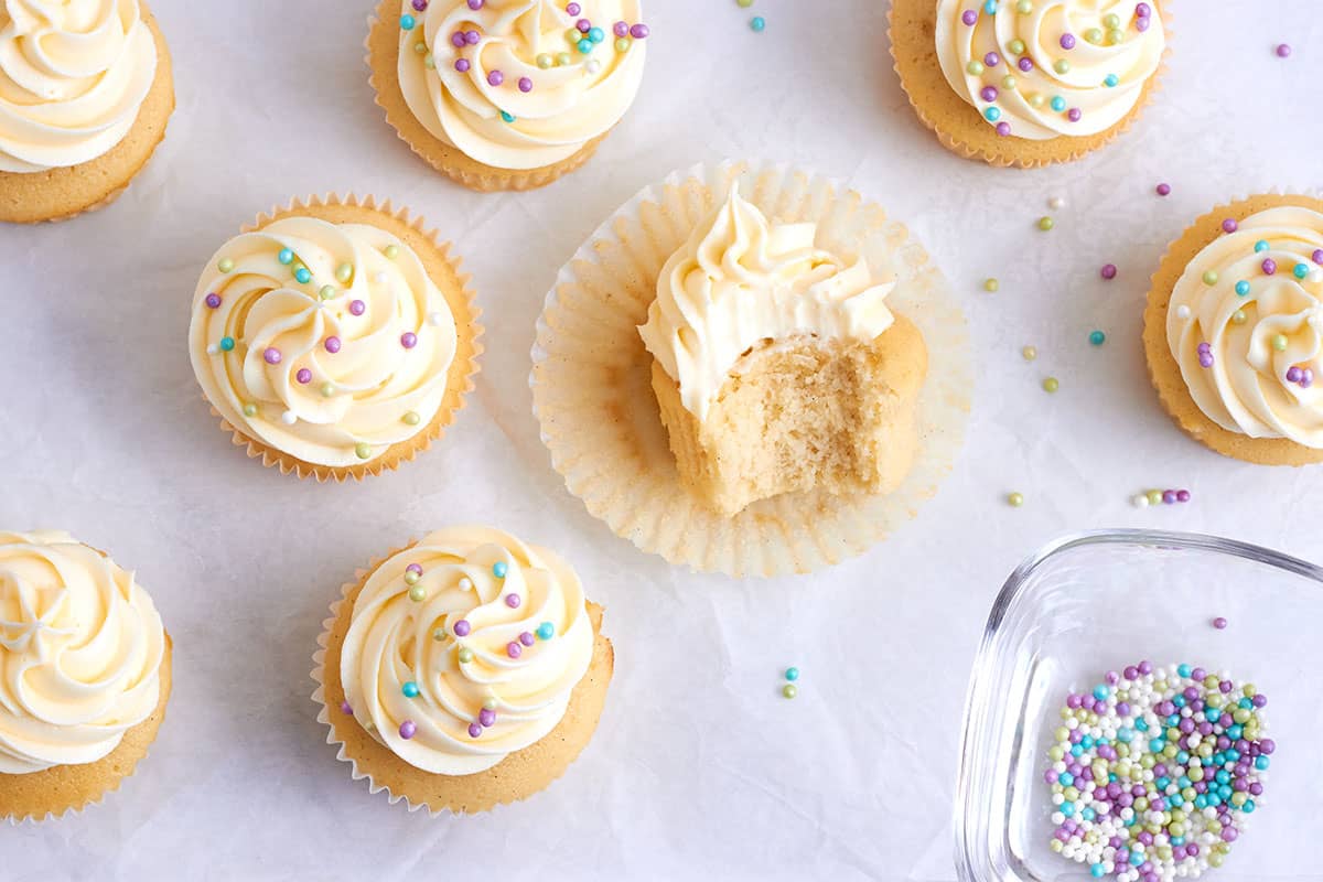 von oben: mehrere vanille cupcakes mit weißem Topping und pastellfarbenden streuseln, in der Mitte ein cupcake von dem ein Biss fehlt