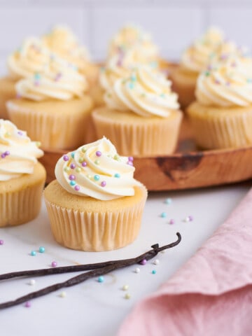 vanilla cupcakes topped with white frosting arranged on a wooden platter and in the foreground