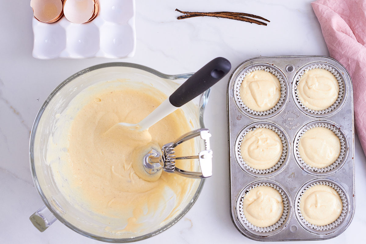 glass mixing bowl filled with creamy cupcake batter, to the right 6 moulds with papers filled with batter